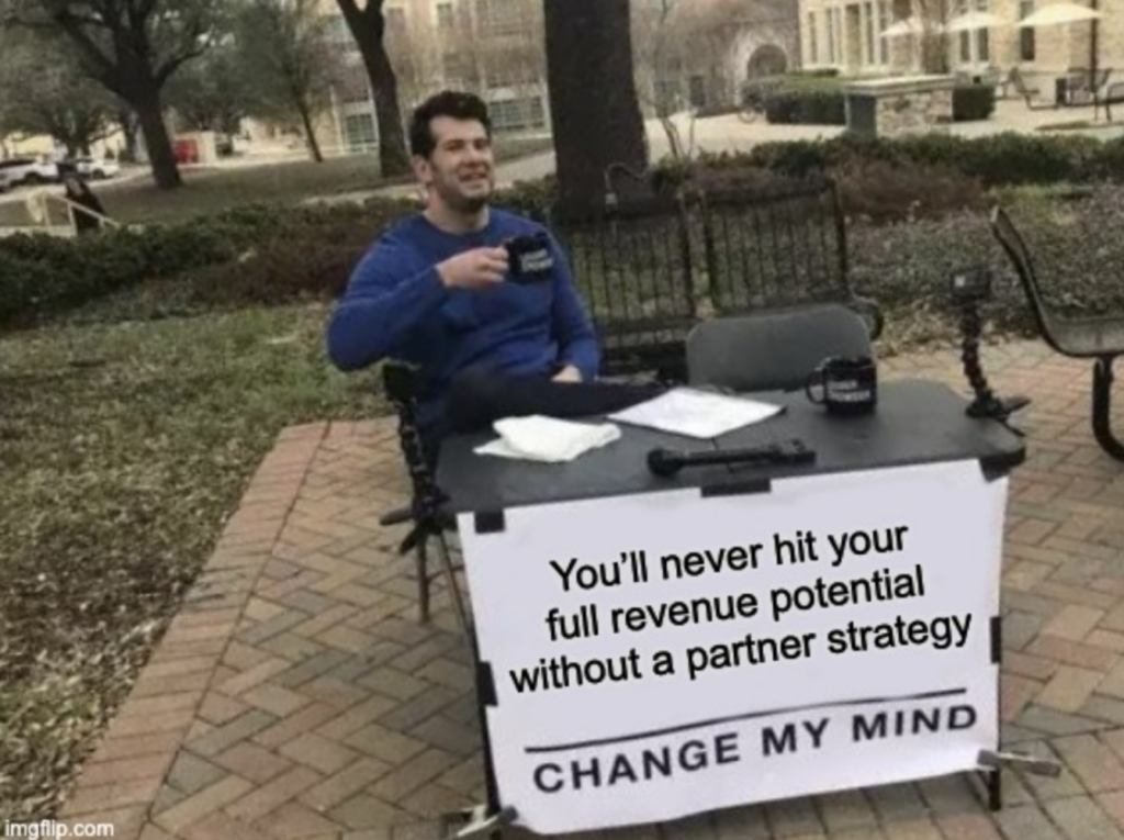 Man at a table with a sign that says "You'll never hit your full revenue potential without a partner strategy. CHANGE MY MIND."